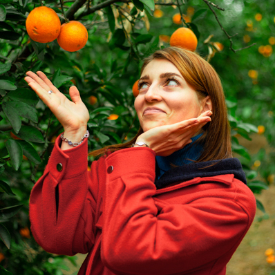 Girl in red coat in garden putting her hands around oranges hanging from orange tree.