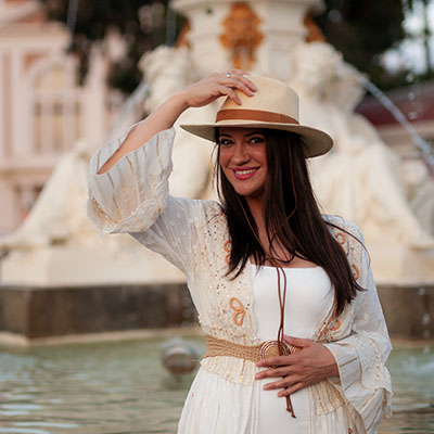 Smiling girl in a beige hat in beautiful white dress standing next to the fountain in Spain