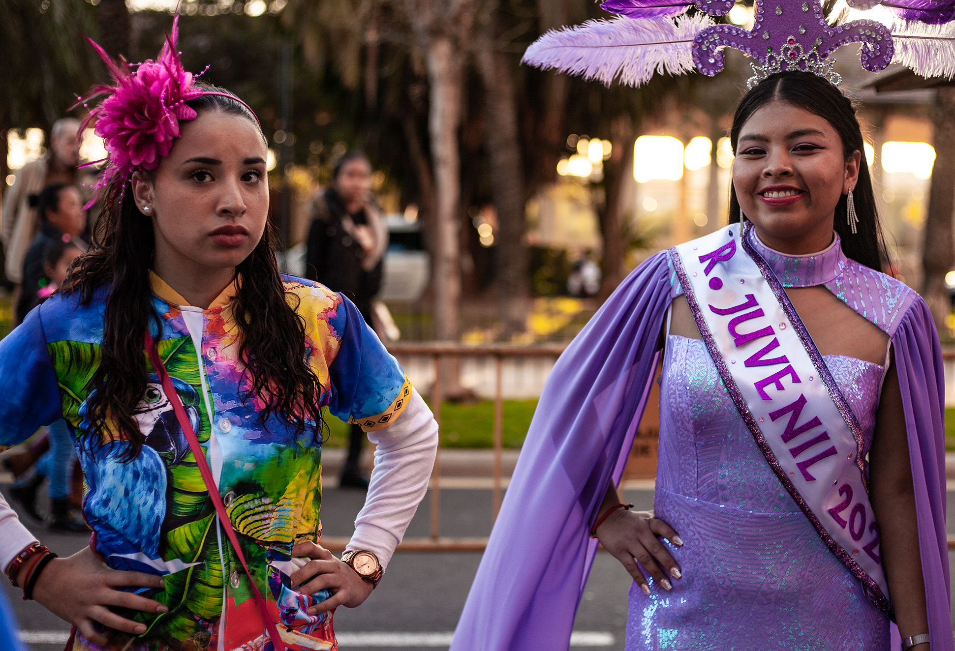 Two gilrs, one with sad look with colorful dress with painting of a blue parrot head, other girl smilling dressed in a flossing purple dress