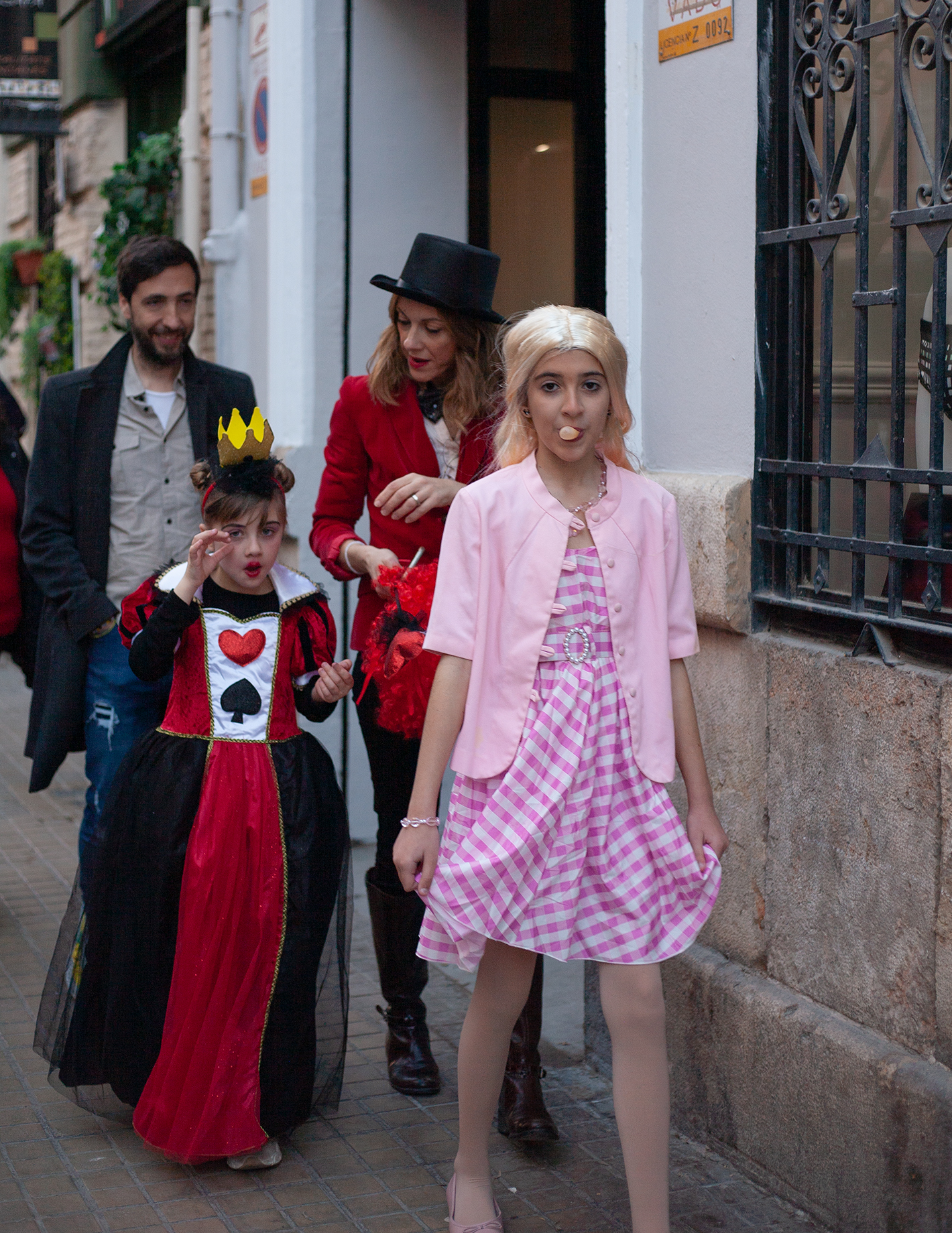 Family of 4 walking the street, mother wearing a dark read blazor and black cylinder, in front teenage girl in pink checked dress and pink blazor and popping a gum ballon