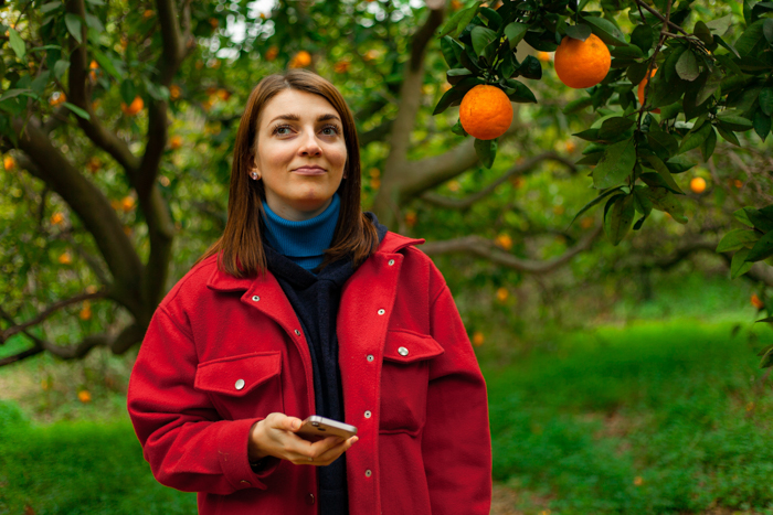 Girl in red coat, brown hair posing in garden with orange trees in background.