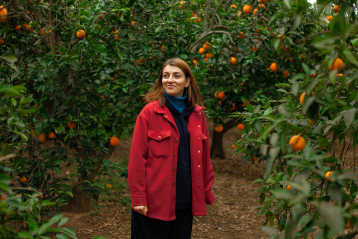 Girl in red coat, brown hair posing in garden with orange trees in background.