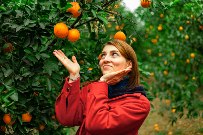 Girl in red coat, brown hair posing in garden with orange trees in background.