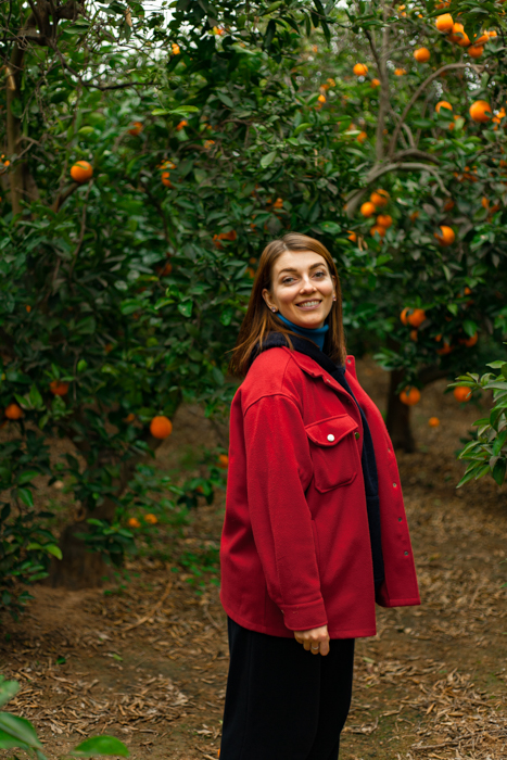 Girl in red coat, brown hair posing in garden with orange trees in background.