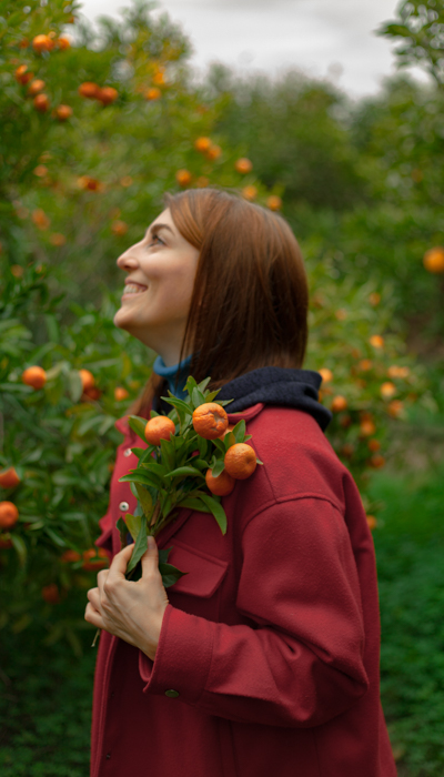 Girl in red coat, brown hair posing in garden with orange trees in background.