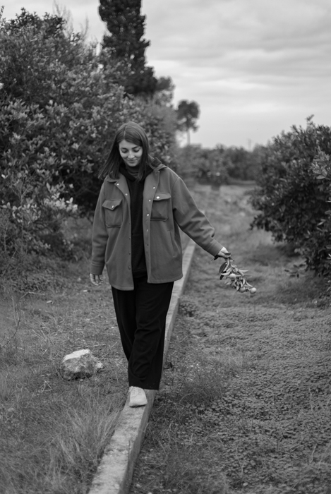 Girl in red coat, brown hair posing in garden with orange trees in background.