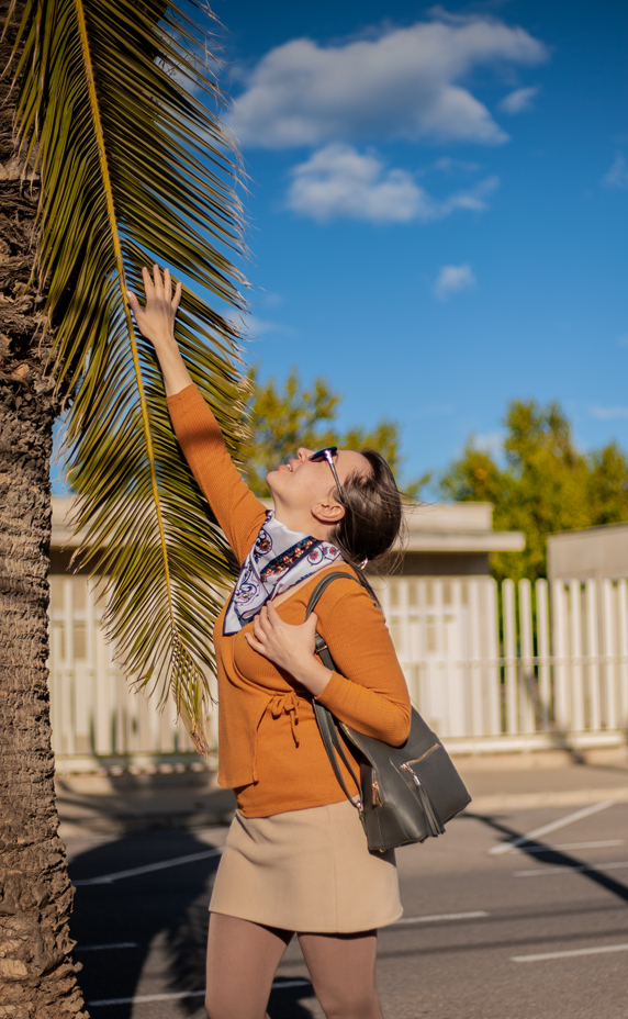 Girl in red coat, brown hair posing in garden with orange trees in background.