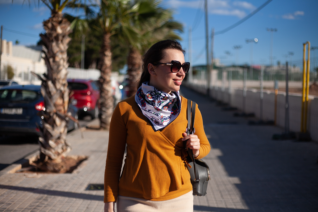 Girl in red coat, brown hair posing in garden with orange trees in background.